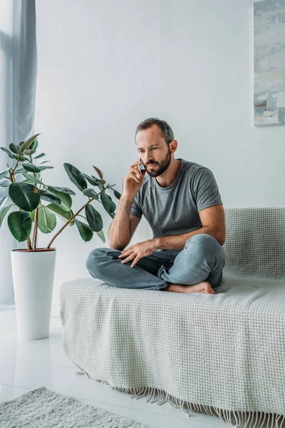 Unhappy bearded man sitting on couch and talking by smartphone — Stock Photo