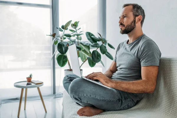 Seitenansicht eines nachdenklichen bärtigen Mannes, der auf der Couch sitzt und Laptop benutzt — Stockfoto