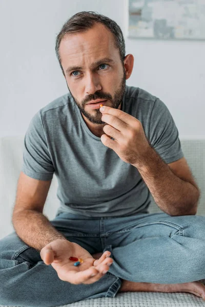 Hombre barbudo infeliz sentado en el sofá y tomando pastillas - foto de stock