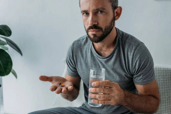 Homem de meia idade barbudo olhando para a câmera enquanto segurando pílulas e vidro de água — Fotografia de Stock