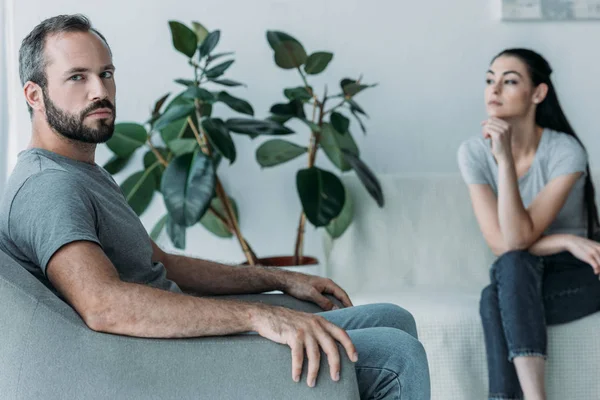 Sconvolto uomo barbuto seduto sulla poltrona e guardando la fotocamera mentre la donna frustrato seduto sul divano e guardando altrove, concetto di difficoltà di relazione — Foto stock