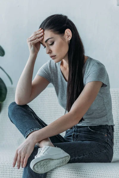 Расстроенная молодая женщина с рукой на лбу сидит на диване и смотрит вниз — Stock Photo