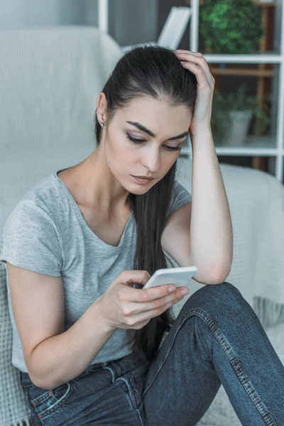 Malheureuse jeune femme assise sur le canapé et utilisant un smartphone — Photo de stock