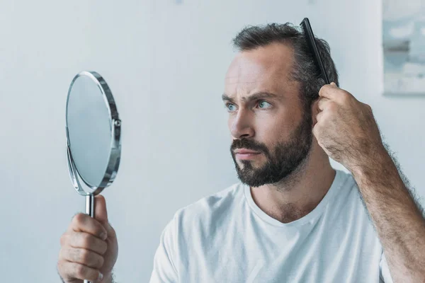 Бородатый мужчина средних лет расчесывает волосы и смотрит на зеркало, концепция выпадения волос — стоковое фото