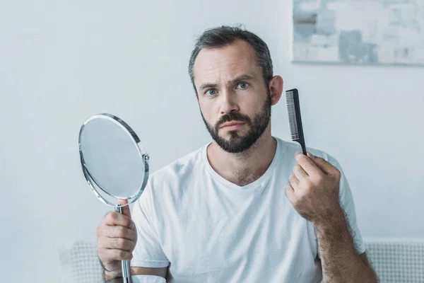 Hombre de mediana edad sosteniendo espejo con peine y mirando a la cámara, concepto de pérdida de cabello - foto de stock