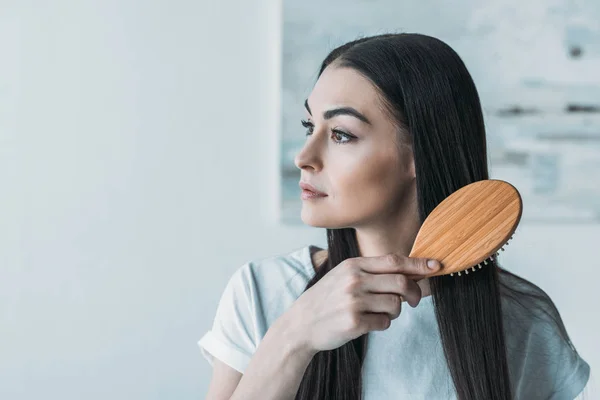 Junge brünette Frau kämmt Haare mit Haarbürste und schaut weg — Stockfoto