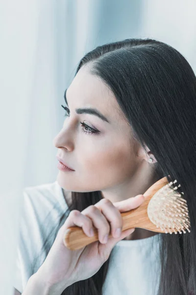 Jovem morena pensiva pentear o cabelo com escova de cabelo e olhando para longe — Fotografia de Stock