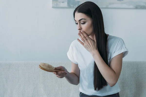 Schockierte brünette Frau sitzt auf der Couch und schaut auf Haarbürste, Haarausfall Konzept — Stockfoto