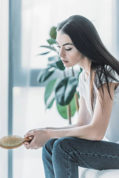 Side view of young sad woman holding hairbrush while sitting indoors, hair loss concept — Stock Photo
