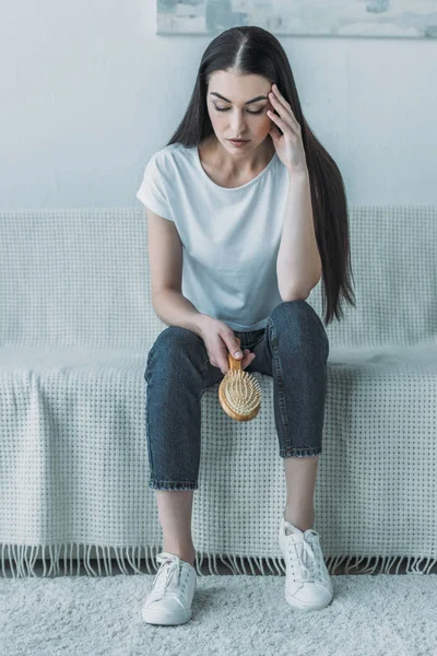 Sconvolto giovane donna tenendo spazzola per capelli e seduto sul divano, concetto di perdita di capelli — Foto stock