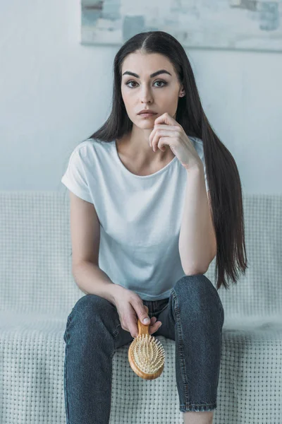 Junge brünette Frau mit Haarbürste, während sie auf der Couch sitzt und in die Kamera schaut — Stockfoto