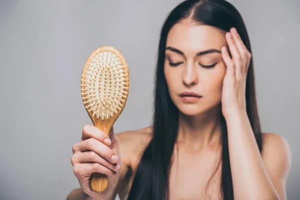 Stressed brunette woman with closed eyes holding hairbrush isolated on grey, hair loss concept — Stock Photo