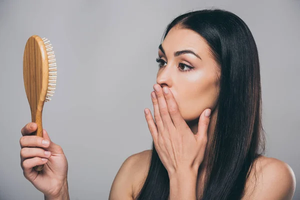Schockierte junge brünette Frau schaut auf Haarbürste isoliert auf grau, Haarausfall Konzept — Stockfoto