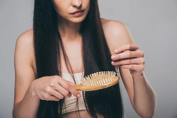 Обрезанный снимок молодой брюнетки расчесывающей волосы расческой, изолированной на сером, концепция выпадения волос — стоковое фото
