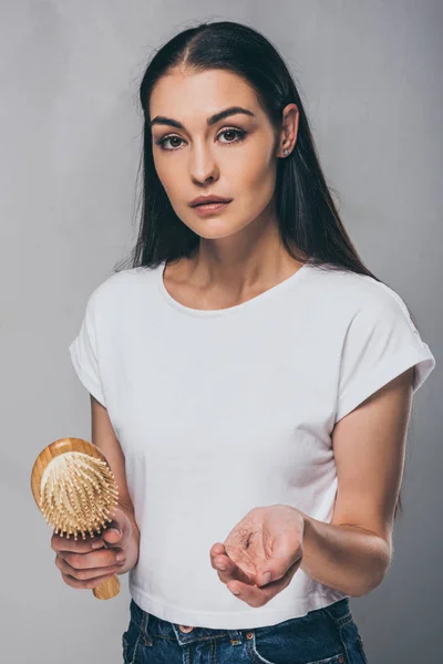 Jeune femme tenant une brosse à cheveux avec les cheveux tombés et regardant la caméra isolée sur gris — Photo de stock