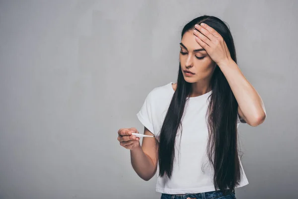 Mujer joven enferma con la mano en la frente mirando el termómetro aislado en gris — Stock Photo