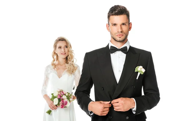 Handsome young groom buttoning jacket and looking at camera with bride standing blurred on background isolated on white — Stock Photo