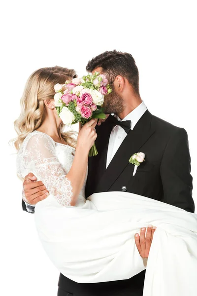 Young newlyweds covering faces with bridal bouquet while groom carrying bride isolated on white — Stock Photo