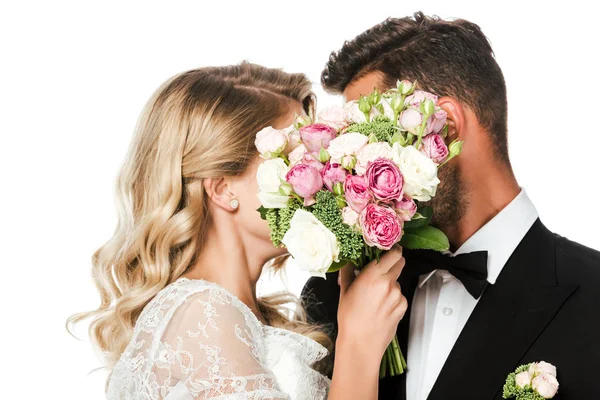 Young bride and groom covering faces with bridal bouquet isolated on white — Stock Photo