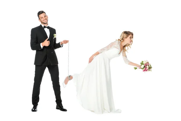 Feliz novio celebración cadena atado alrededor novias pierna y mostrando pulgar hacia arriba aislado en blanco - foto de stock