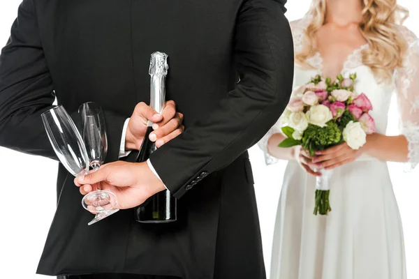 Schnappschuss des Bräutigams mit Champagnerflasche und Gläsern hinter dem Rücken, während die Braut isoliert auf weiß vor ihm steht — Stockfoto