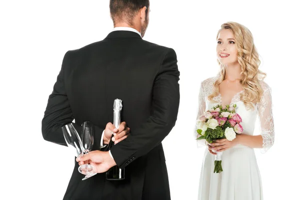 Vue arrière du marié tenant bouteille de champagne et lunettes derrière le dos tandis que la mariée debout devant lui isolé sur blanc — Photo de stock