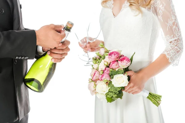 Cropped shot of groom opening champagne bottle while bride holding glasses isolated on white — Stock Photo