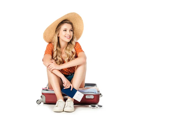 Feliz joven en sombrero de paja con pasaporte y billete sentado en la maleta aislado en blanco - foto de stock