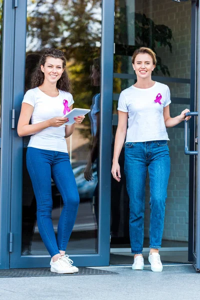 Women with pink breast cancer awareness ribbons smiling at camera near modern building — Stock Photo