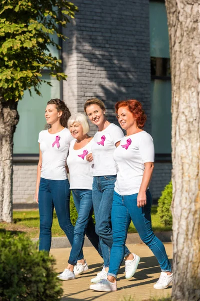 Mulheres sorridentes com fitas de conscientização sobre câncer de mama de mãos dadas e caminhando juntas — Fotografia de Stock