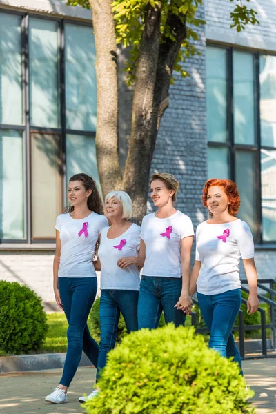 Mujeres felices con cintas de conciencia sobre el cáncer de mama cogidas de la mano y caminando juntas - foto de stock