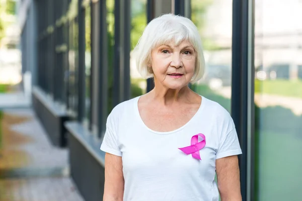 Mujer mayor con cinta rosa en la camiseta mirando a la cámara, concepto de conciencia de cáncer de mama - foto de stock