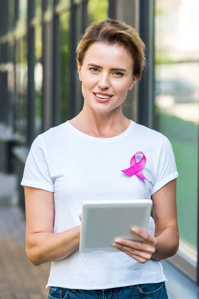 Woman with pink ribbon on t-shirt using digital tablet and smiling at camera, breast cancer awareness concept — Stock Photo