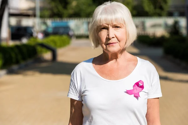 Mulher idosa com fita rosa em pé na rua e olhando para a câmera, conceito de consciência do câncer de mama — Fotografia de Stock