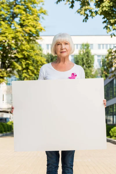 Пожилая женщина с розовой лентой, держащая пустой баннер и глядя на камеру, концепция рака молочной железы — стоковое фото