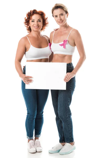 Mujeres en sostenes con cintas de conciencia de cáncer de mama sosteniendo la tarjeta en blanco y sonriendo a la cámara aislada en blanco - foto de stock