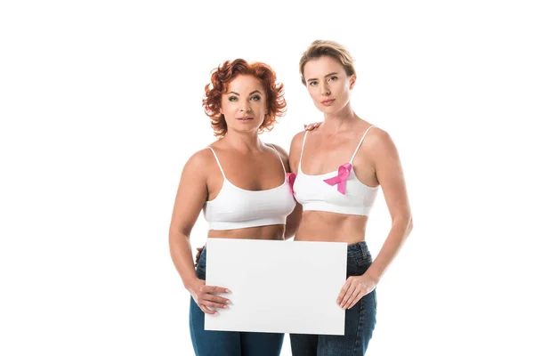 Mujeres en sostenes con cintas de conciencia de cáncer de mama sosteniendo la tarjeta en blanco y mirando a la cámara aislada en blanco - foto de stock