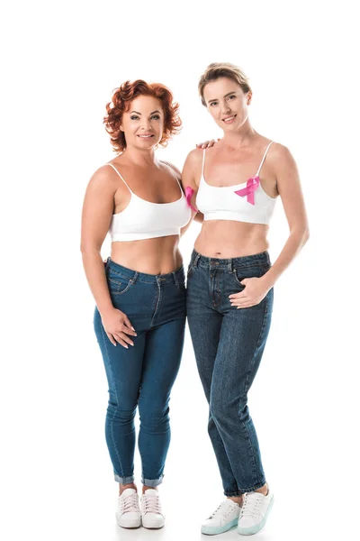 Women in bras with breast cancer awareness ribbons standing together and smiling at camera isolated on white — Stock Photo