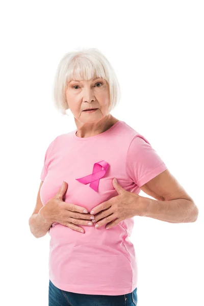 Mujer mayor con cinta rosa tocando el pecho y mirando a la cámara aislada en blanco, concepto de cáncer - foto de stock
