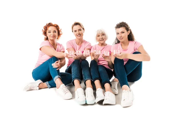 Femmes en t-shirts roses tenant des cubes avec le mot cancer et souriant à la caméra tout en étant assis ensemble isolé sur blanc — Stock Photo