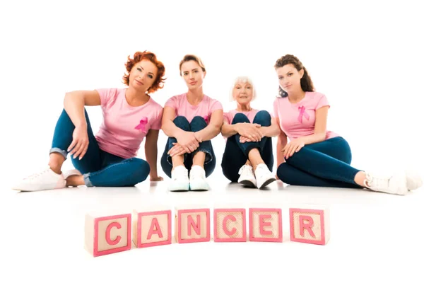 Femmes en t-shirts roses assis et regardant la caméra, mot cancer isolé sur blanc — Photo de stock