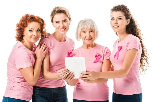 Donne in t-shirt rosa con nastri di consapevolezza del cancro al seno che tengono tablet digitale e sorridono alla fotocamera isolata su bianco — Foto stock