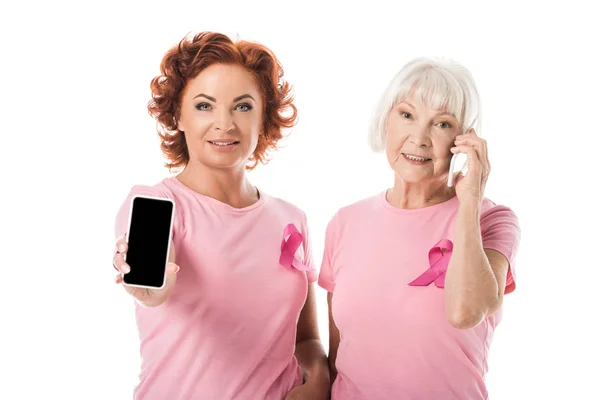 Donne con nastri rosa utilizzando smartphone e sorridendo alla fotocamera isolata sul bianco, concetto di consapevolezza del cancro al seno — Foto stock