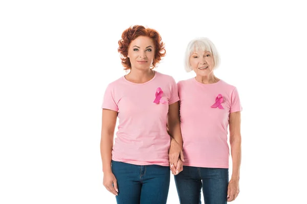 Mature and senior women with pink ribbons holding hands and smiling at camera isolated on white, breast cancer awareness concept — Stock Photo