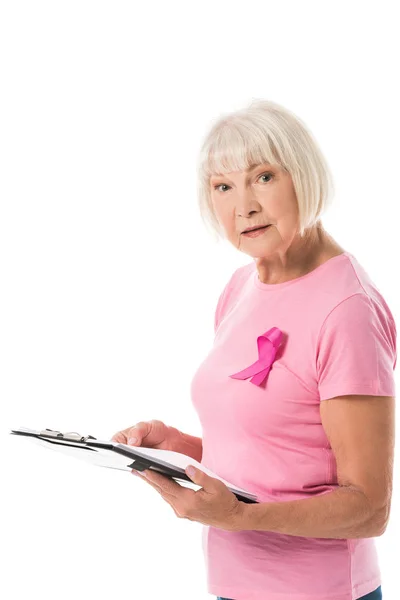 Femme âgée en t-shirt rose avec ruban de sensibilisation au cancer du sein tenant presse-papiers et regardant la caméra isolée sur blanc — Photo de stock