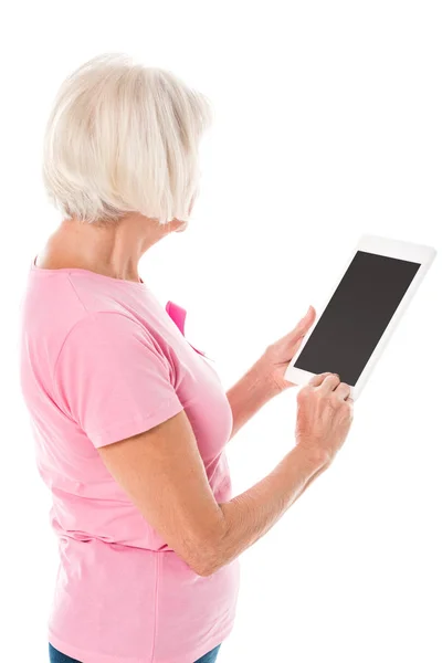 Femme âgée en t-shirt rose avec ruban de sensibilisation au cancer du sein à l'aide d'une tablette numérique avec écran blanc isolé sur blanc — Photo de stock