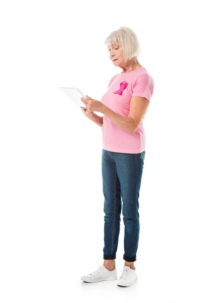 Femme âgée en t-shirt rose avec ruban de sensibilisation au cancer du sein en utilisant une tablette numérique isolée sur blanc — Photo de stock