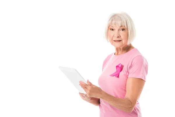 Femme âgée en t-shirt rose avec ruban de sensibilisation au cancer du sein en utilisant une tablette numérique et en regardant la caméra isolée sur blanc — Photo de stock