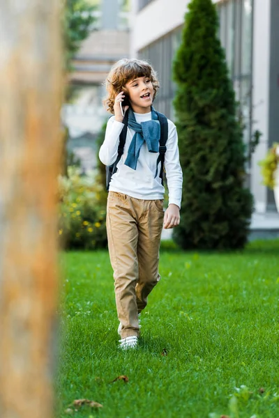 Enfoque selectivo de niño sonriente con mochila hablando por teléfono inteligente y caminando sobre césped verde - foto de stock