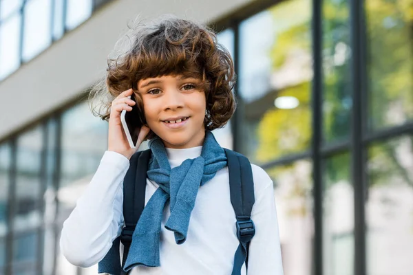 Cute preteen schoolboy with backpack talking by smartphone and smiling at camera — Stock Photo